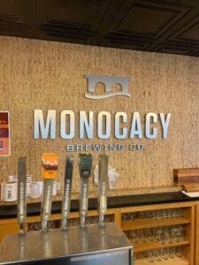 A wall with the large silver letters “MONOCACY BREWING CO” and a bridge logo rises above a modern bar counter. Five taps labeled with “Monocacy Brewing Co.” hint at why it’s among the best breweries in Frederick MD. Shelves below hold clean glassware.