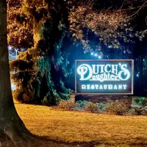 A brightly lit sign at night reads Dutch’s Daughter Restaurant, one of the best bars in Frederick. The sign is surrounded by greenery and trees, with fallen leaves covering the ground and a warm glow shining against the dark, wooded background.