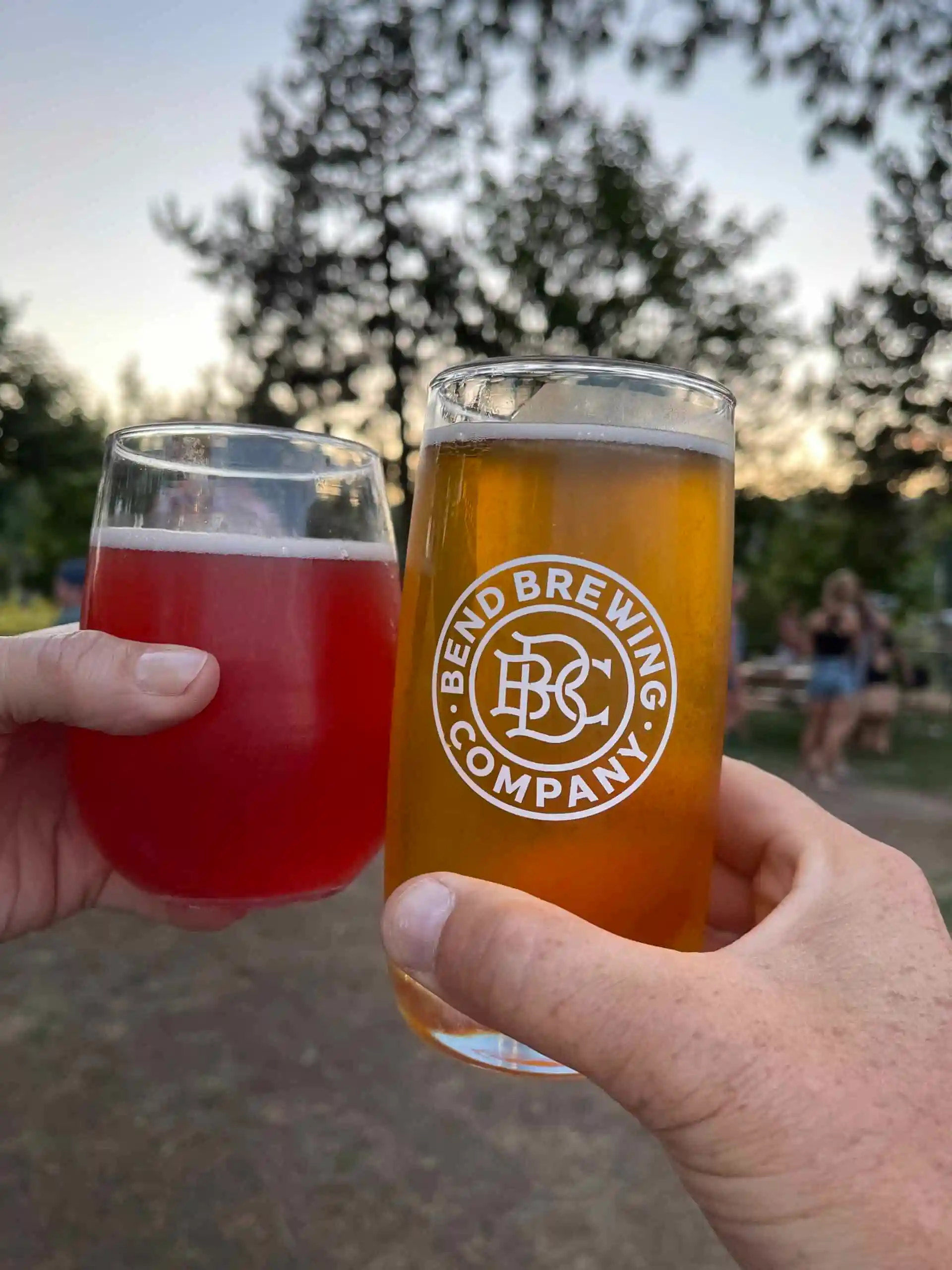 Two Hands Clink Glasses Outdoors One Holds a Red Drink in a Stemless Wine Glass the Other a Pint of Amber Beer with the Bend Brewing Company Logo Trees and People Blur in the Background Evoking a Casual Dusk Gathering in Bend Oregon