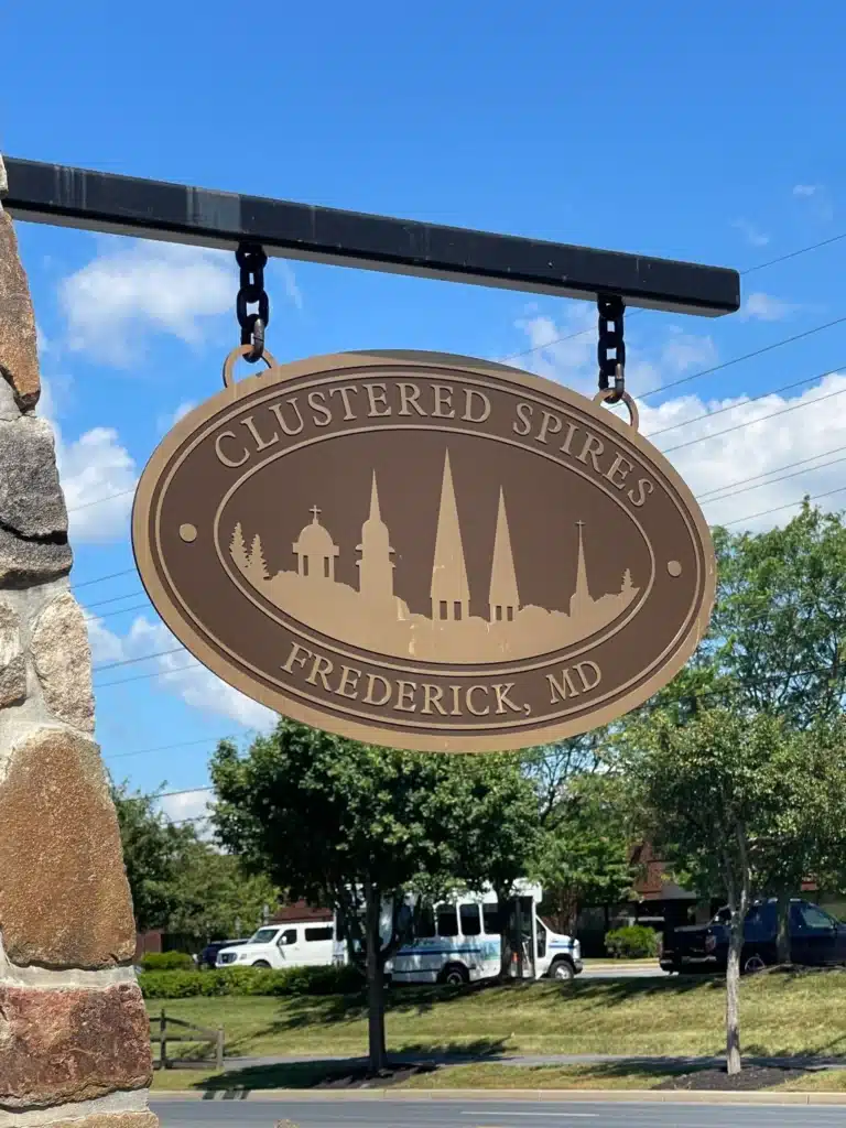 Oval Tan Sign Reading Clustered Spires Frederick Md Featuring Church Spires Silhouettes Hangs from a Metal Bracket on a Stone Pillar in the Background Are Trees Parked White Vans and a Blue Sky with Clouds Near New Market Md
