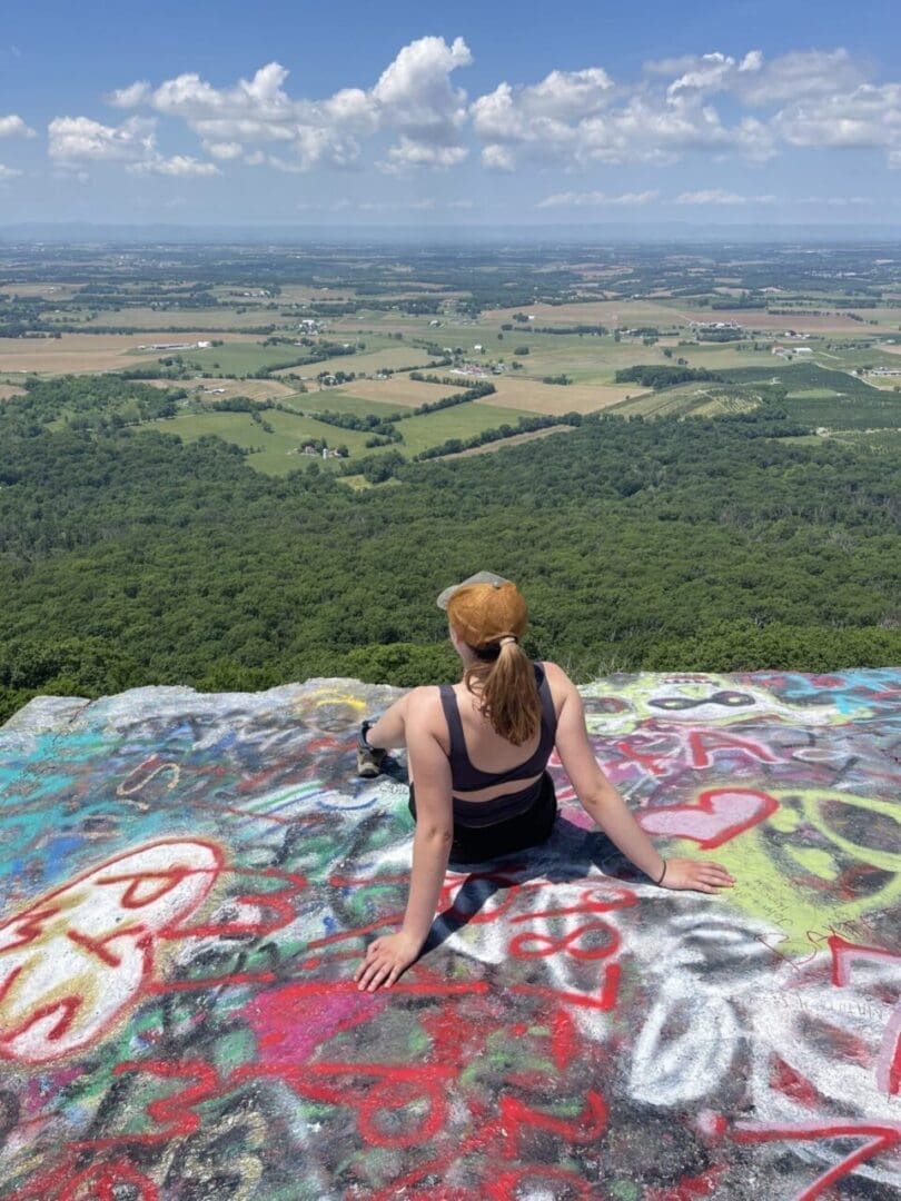 a Woman with a Ponytail and Tan Cap Sits on a Colorful Graffiti Covered Rock Ledge Enjoying One of the Best Hikes Around Frederick Md As She Overlooks Green Fields and Forests Under a Bright Blue Sky with Scattered Clouds