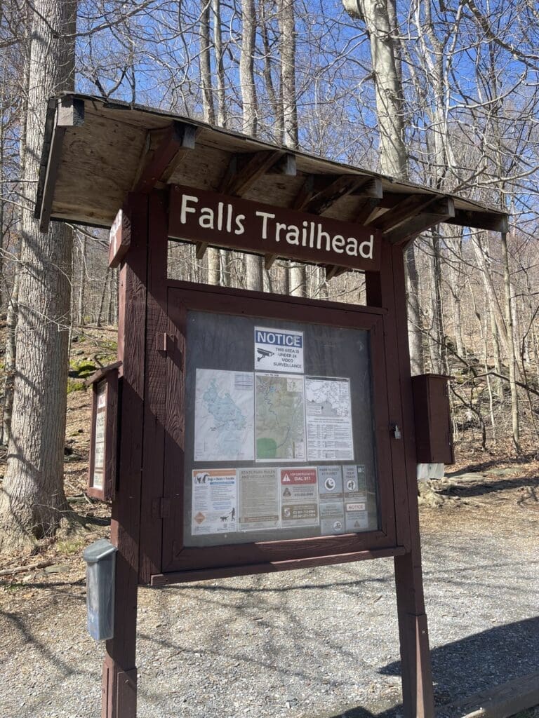 a Wooden Trailhead Sign Labeled Falls Trailhead Stands in a Forest with Bare Treesone of the Best Hikes Around Frederick Md the Sign Displays Maps Notices and Safety Info Inside a Glass Case As Sunlight Highlights the Gravel Covered Ground