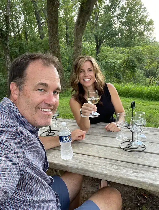 A smiling man and woman sit at an outdoor wooden picnic table in a green, wooded area, enjoying drinks from Frederick MD Wineries. The man holds a water bottle; the woman sips white wine. Empty wine glass holders rest on the table.