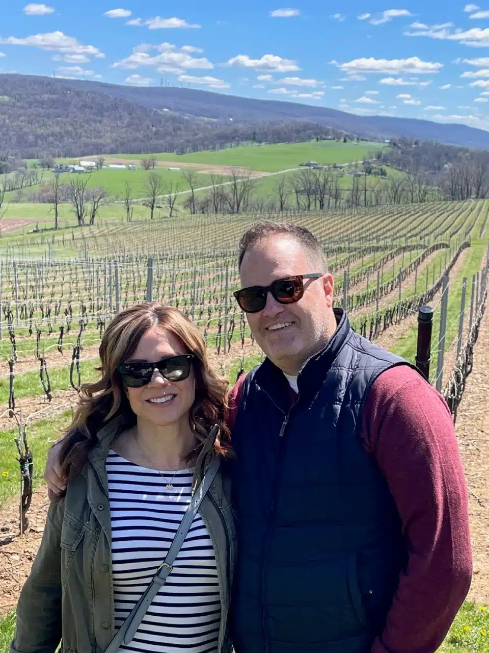 A woman and man wearing sunglasses stand smiling in front of a vineyard at one of the Frederick MD Wineries, with rows of bare grapevines, green hills, scattered trees, and a blue sky dotted with white clouds in the background.
