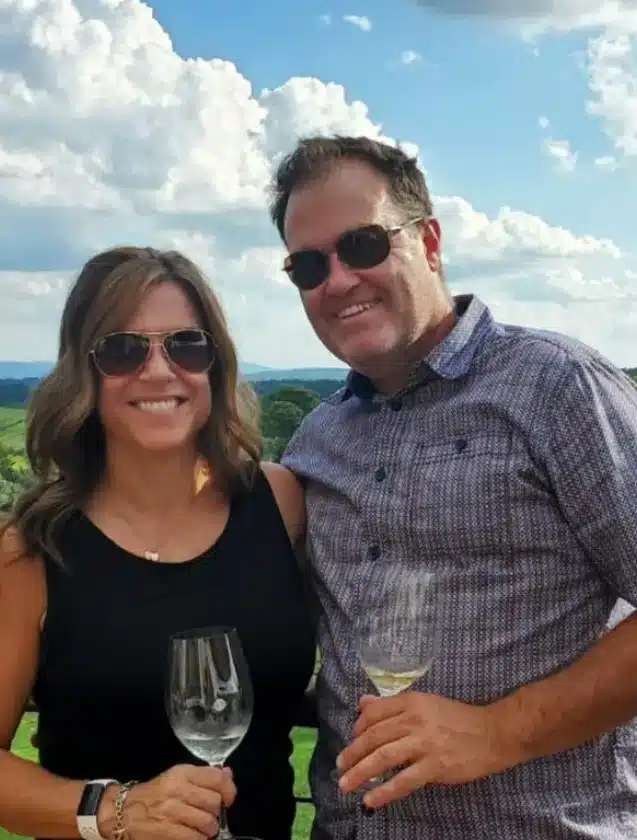 A smiling man and woman wearing sunglasses stand outdoors at one of the Frederick MD Wineries, holding wine glasses. The woman has long brown hair and a sleeveless black top; the man wears a patterned shirt. Green landscape and blue sky complete the scene.