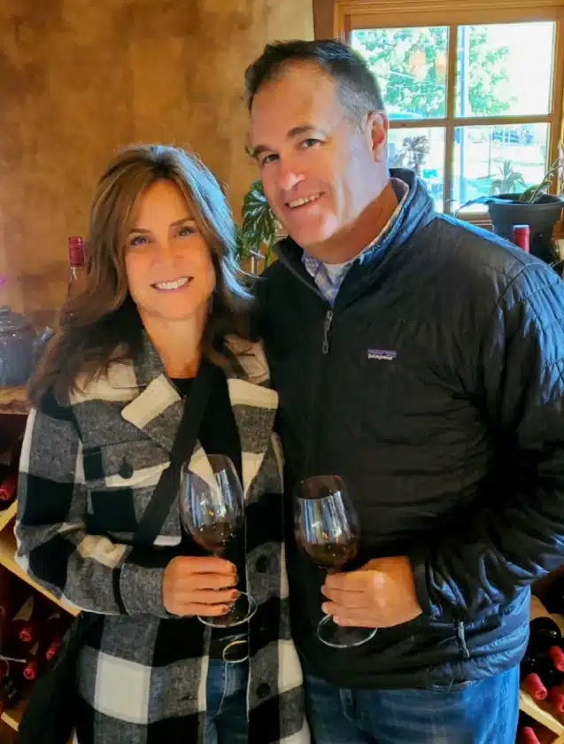 A smiling woman and man stand close together indoors at one of the Frederick MD Wineries, each holding a glass of red wine. The woman wears a plaid coat and the man wears a dark jacket. Behind them are shelves with wine bottles and a window with greenery outside.