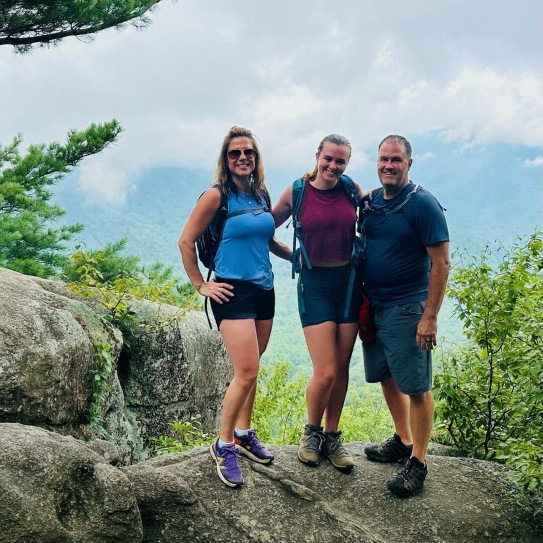 Three People Two Women and One Man Stand Smiling on a Large Rock Surrounded by Green Trees Ready to Explore Some of the Best Hikes Around Frederick Md a Misty Mountainous Landscape Appears in the Background Under a Cloudy Sky