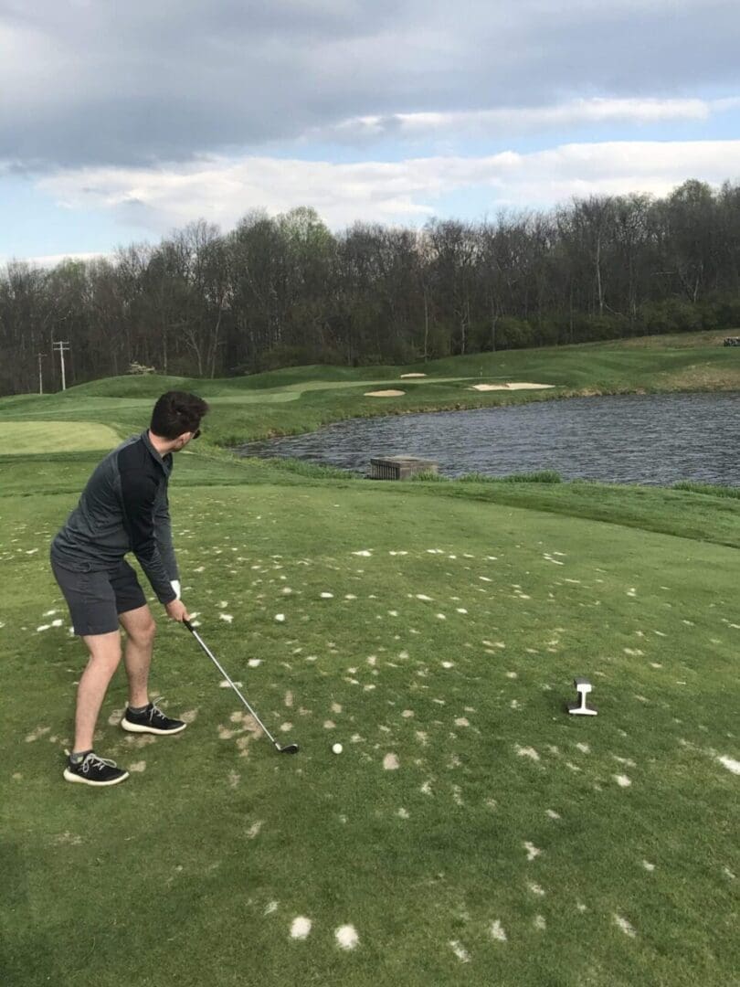 Whiskey creek golf par three a Man in a Gray Long sleeve Shirt and Shorts Prepares to Swing a Golf Club on a Patchy Green Fairway Near a Pond Typical of Scenic Golf Courses Near Frederick Md with Trees and Sand Traps in the Background Under a Partly Cloudy Sky