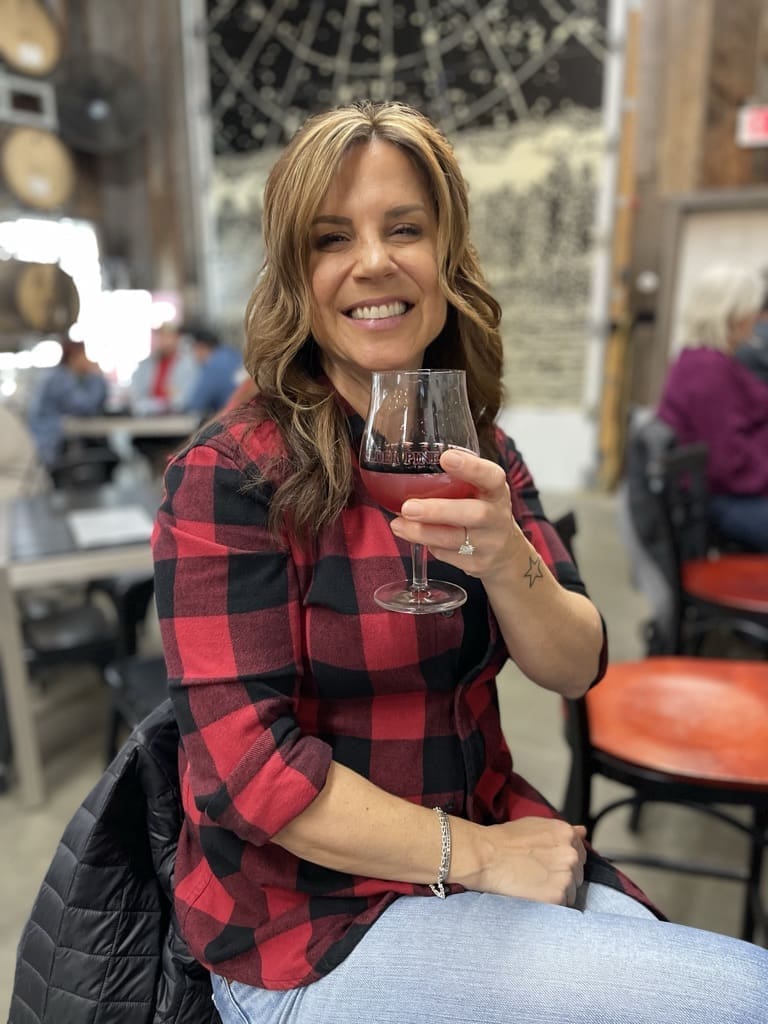 Nicole elder pine brewery a Woman with Wavy Brown Hair Wearing a Red and Black Plaid Shirt and Jeans Smiles While Holding a Glass of Red Wine She Sits Indoors at a Table in One of the Best Breweries in Frederick Md with Barrels and People Blurred in the Background