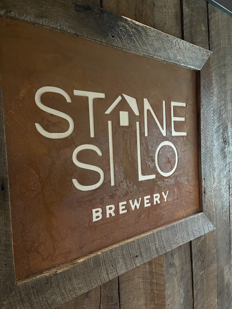 a Rustic Wooden Sign Displays the Text Stone Silo Brewery in White with the a in Stone Styled As a Roof with a Small Chimney to Resemble a House Set Against a Weathered Rusty Orange Background and Rough Wooden Frame It Captures New Market Md Charm