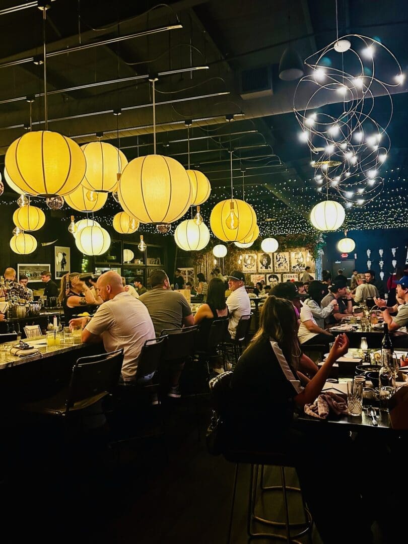 a Lively Bend Restaurants Bar Scene with People Seated at Tables and the Counter Warmly Lit by Large Yellow Globe Lanterns and String Lights Overhead the Background Features Framed Art on Dark Walls and Bartenders Serving Drinks