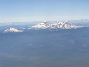 Aerial view of a mountain range with snow-capped peaks under a clear blue sky, dense forests below. This stunning landscape showcases the natural beauty and things to do in Bend OR, with mountains rising above light haze near the ground.