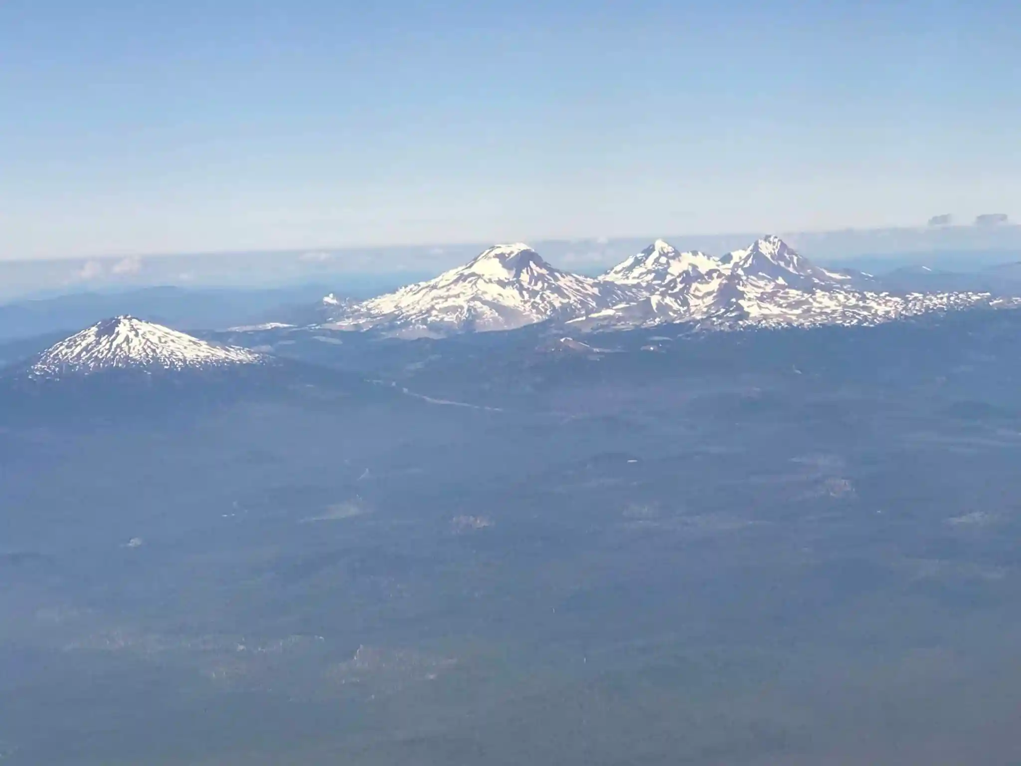 Aerial View of a Mountain Range with Snow Capped Peaks Under a Clear Blue Sky Dense Forests Below This Stunning Landscape Showcases the Natural Beauty and Things to Do in Bend or with Mountains Rising Above Light Haze Near the Ground