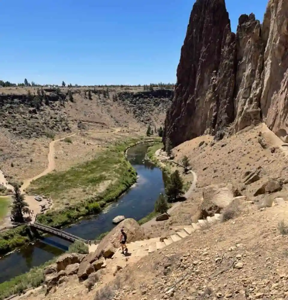 a Person Climbs Stone Steps Along a Dry Hillside Under a Clear Blue Skya Classic Scene from Hikes Near Bend Oregon Below a Winding River Flows Past Green Grass While Tall Jagged Rock Formations and Walking Paths Define the Stunning Landscape