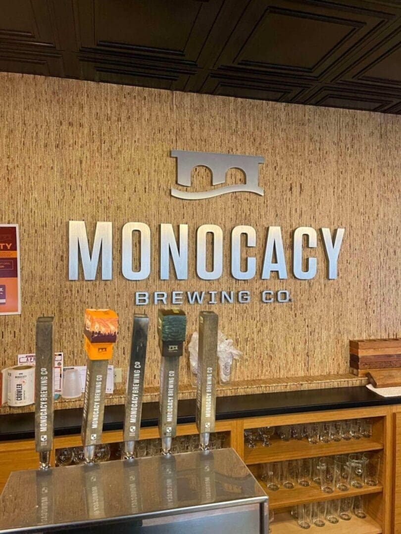 a Wall with the Large Silver Letters Monocacy Brewing Co and a Bridge Logo Rises Above a Modern Bar Counter Five Taps Labeled with Monocacy Brewing Co Hint at Why Its Among the Best Breweries in Frederick Md Shelves Below Hold Clean Glassware