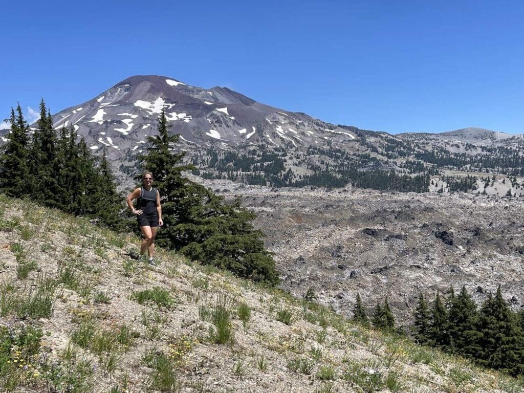 a Woman in Athletic Clothing Hikes Up a Rocky Grassy Slope Near Bend Orone of the Top Things to Do in Bend Orsurrounded by Wildflowers and Small Trees with a Snow Patched Mountain Rising Under a Clear Blue Sky