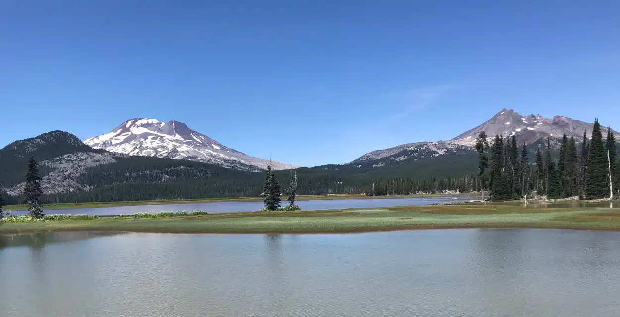 a Calm Lake in the Foreground Reflects a Clear Blue Sky with Patches of Green Grass and Scattered Pine Trees on the Shore in the Background Two Snow Patched Mountains Rise Above Dense Forestperfect for Hikes Near Bend Oregon in a Serene Natural Landscape