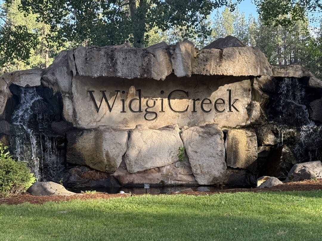 a Large Stone Sign Reading Widgi Creek is Set into a Rock Formation with Small Waterfalls on Both Sides Green Grass and Trees Surround This Bend Oregon Golf Course Landmark As Sunlight Casts Shadows Across the Rocks