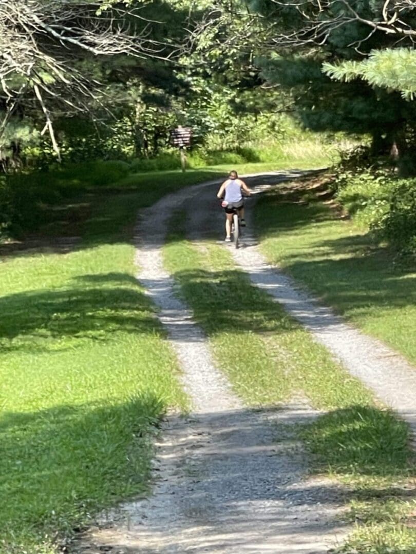 a Person Wearing a Light Colored Tank Top and Dark Shorts Rides a Bicycle Along the Sunlit Little Bennett Bike Trails Where Dappled Sunlight Filters Through Tall Trees and Lush Green Grass Lines the Gravel Path