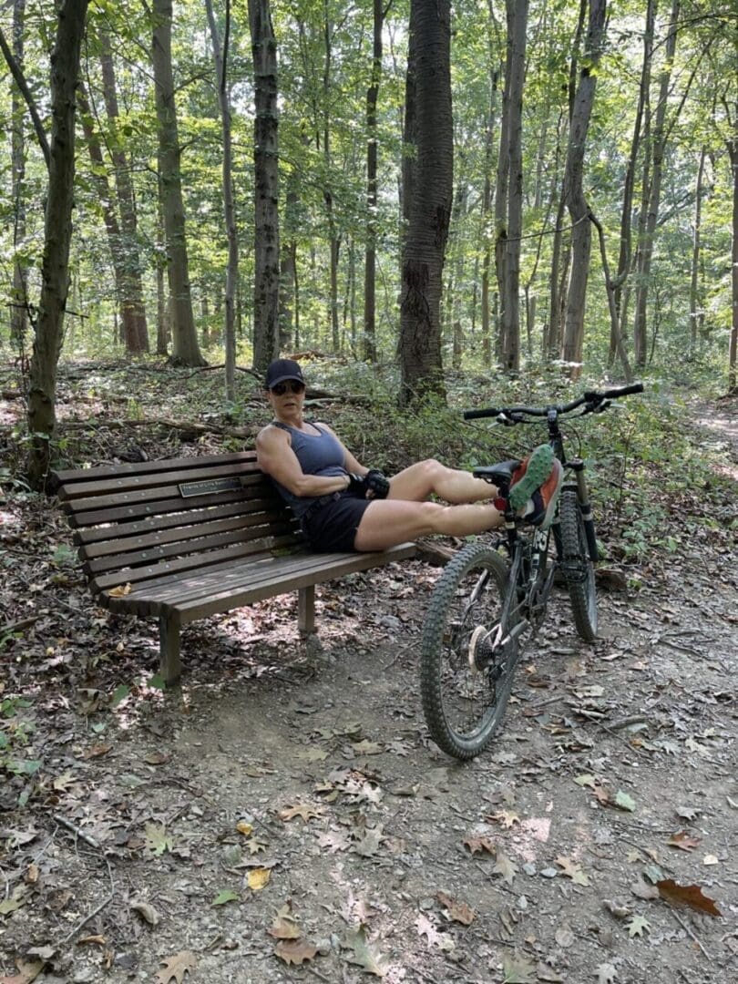 a Person in Athletic Gear Reclines on a Wooden Bench Along the Little Bennett Bike Trails Resting Their Legs on a Mountain Bike They Wear a Cap Sleeveless Shirt Shorts and Sneakers Beneath Tall Trees with Green Foliage and Scattered Leaves