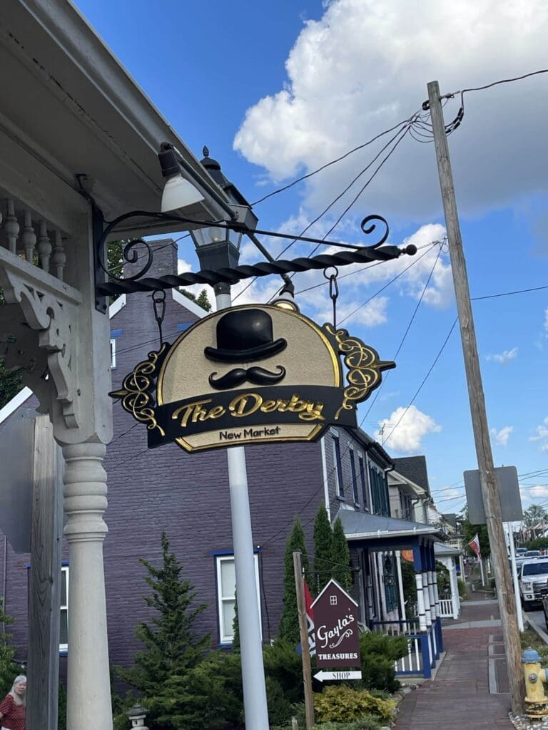 a Decorative Hanging Sign Reads the Derby New Market with a Black Bowler Hat Graphic Above the Text the Sign is Attached to a White Post Near a Brick Building Sidewalk and Blue Skyperfect for Those Exploring Tree Trekkers Frederick Md
