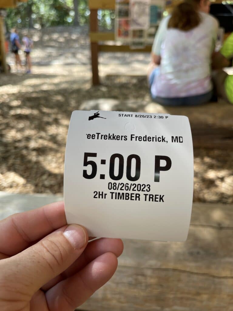 a Hand Holds a White Ticket for Treetrekkers Frederick Md Showing 500 P the Date 08262023 and 2hr Timber Trek in the Blurred Background People Sit at a Wooden Table Outdoors