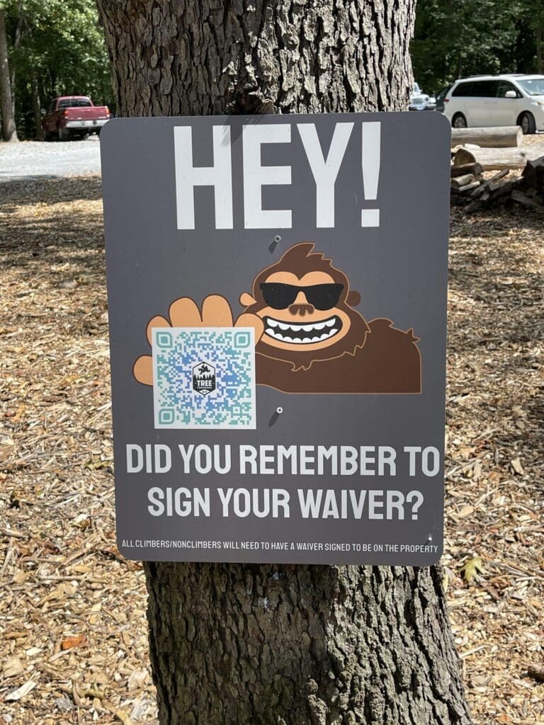 a Sign on a Tree at Tree Trekkers Frederick Md Shows a Cartoon Bigfoot in Sunglasses Waving Text Reads Hey Did You Remember to Sign Your Waiver a Qr Code and Small Print Explain Waiver Requirements for All Climbersnon Climbers Cars and Trees Are Visible Behind