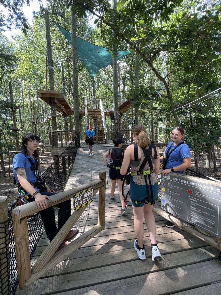 Four People Wearing Harnesses Wait on a Wooden Platform at Tree Trekkers Frederick Md They Face Rope Bridges and Elevated Walkways Among the Trees While Sunlight Filters Through Leaves and an Informational Sign is Visible on the Right