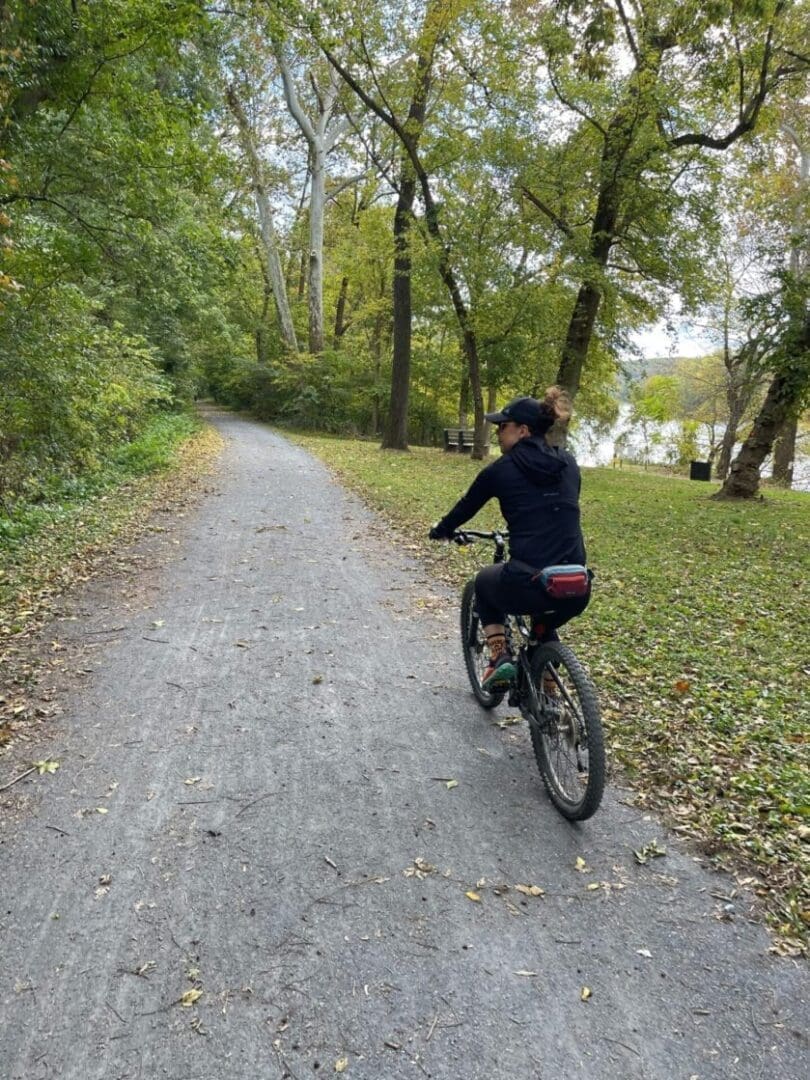 a Person Wearing a Black Outfit and Cap Rides a Bicycle on a Gravel Path Through a Lush Green Forest Along One of the Scenic Bike Trails Near Frederick Md Fallen Leaves Border the Path Trees Line Both Sides and a River is Partially Visible in the Background