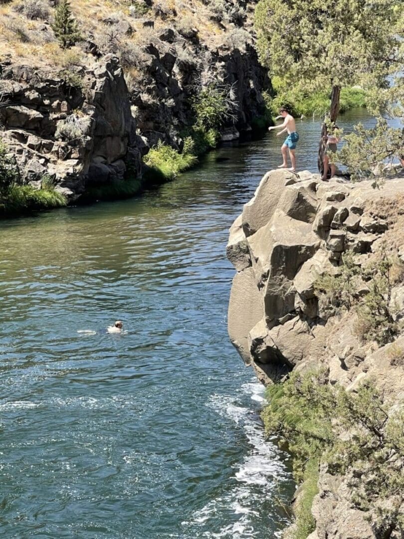 Two People Stand on a Rocky Cliff Above Steelhead Falls One Preparing to Jump into the River Below Another Person Swims Surrounded by Steep Rock Walls and Sparse Trees All Under Bright Sunlight