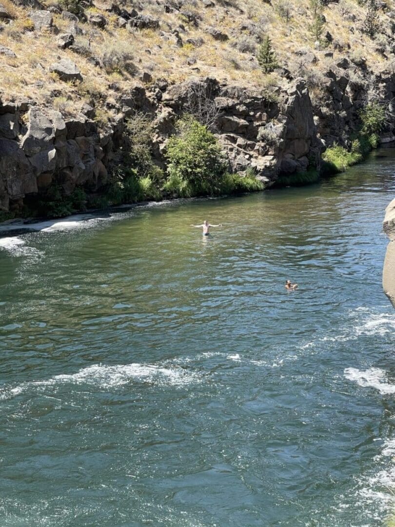 a River with Rippling Blue Green Water Flows Past Rocky Shrub Covered Banks Near Steelhead Falls Two People Are Swimming One in the Center Raising Their Arms As Sunlight Illuminates the Water and Dry Terrain Above the Rocks