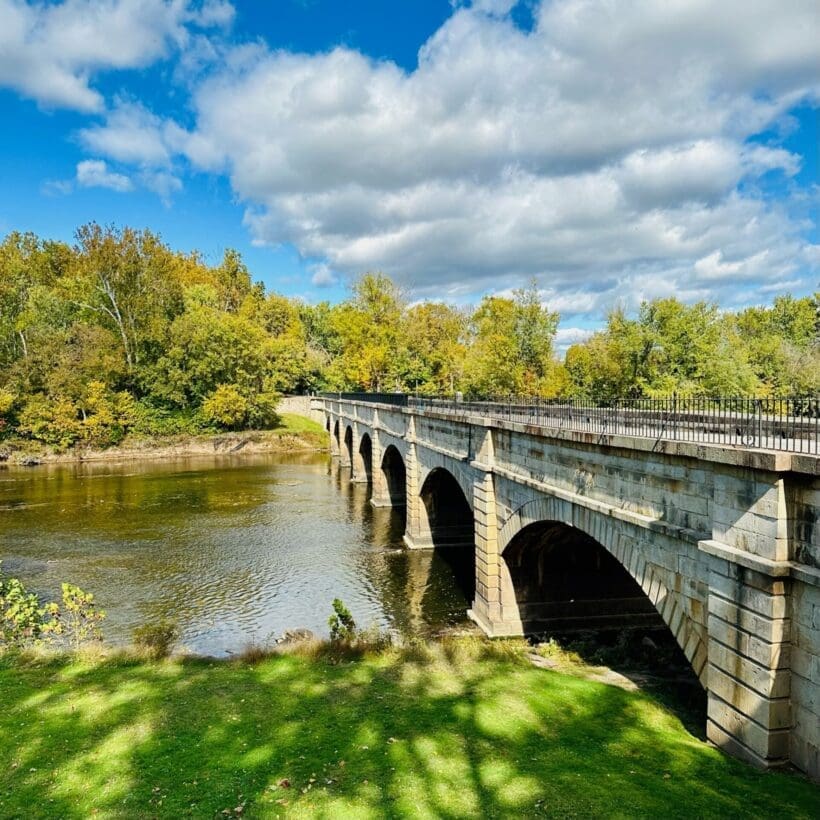 a Stone Arch Bridge Crosses a Calm River Under a Partly Cloudy Sky Lush Green Trees with Hints of Autumn Color Line the Riverbanks While Bike Trails Near Frederick Md Invite Exploration of This Serene Sunlit Landscape