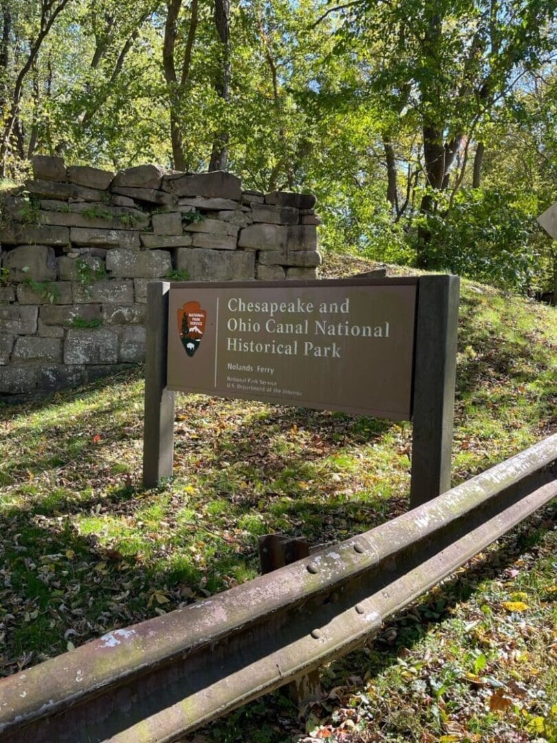 a Brown Sign for Chesapeake and Ohio Canal National Historical Park Stands Amid Grass and Fallen Leaves Near a Wooden Guardrail and Stacked Stone Ruins with Tall Leafy Trees Casting Dappled Sunlightperfect for Exploring Bike Trails Near Frederick Md