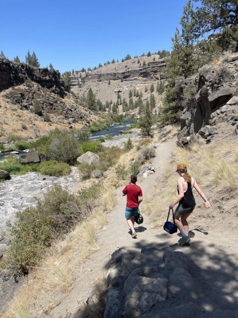 Two People Walk Down a Dusty Trail Beside a River in the Rocky Dry Landscape of Steelhead Falls with Scattered Trees and a Clear Blue Sky One Wears a Red Shirt and Shorts the Other a Tank Top and Shorts Both Carrying Bags