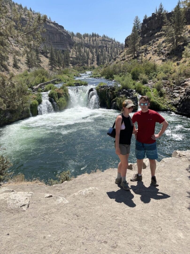 a Woman and Man Stand Side by Side on a Dirt Path Near a Lush River and Waterfall Surrounded by Green Trees and Rocky Hills Both Wear Sunglasses and Casual Hiking Clothes Soaking Up the Sun on One of Many Scenic Bend Hikes