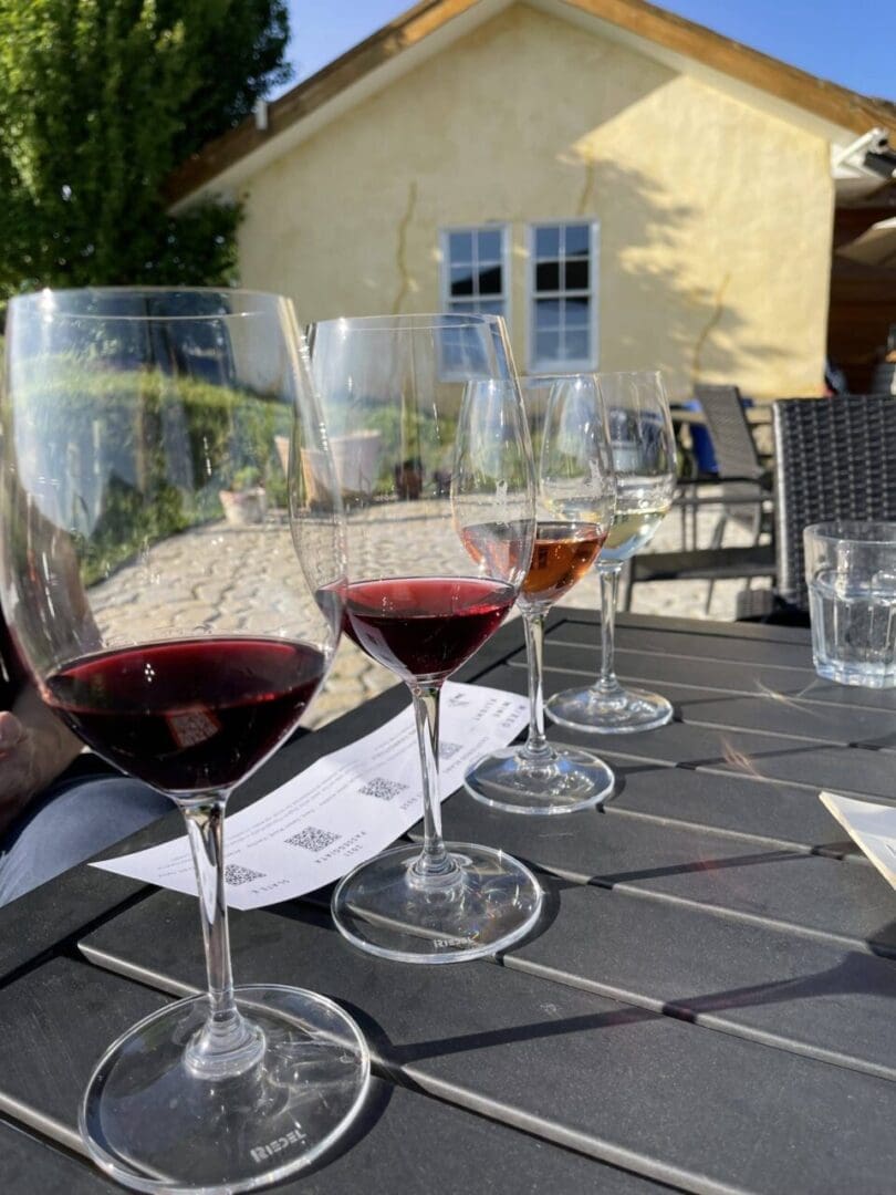 Four Wine Glasses Each with Different Amounts and Colors of Wine Are Arranged on a Black Outdoor Table at Burnt Hill Farm a Glass of Water and a Paper with Qr Codes Sit Nearby with a Yellow House Sunlight and Greenery in the Background