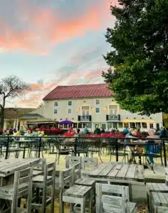 Outdoor Dining Area with Wooden Tables and Chairs Some Occupied by People Enjoying Meals and Conversation Nearby Greenery and a Large Beige Building Set the Scene Beneath a Colorful Sunsetperfect for Relaxing After Visiting Frederick Md Wineries