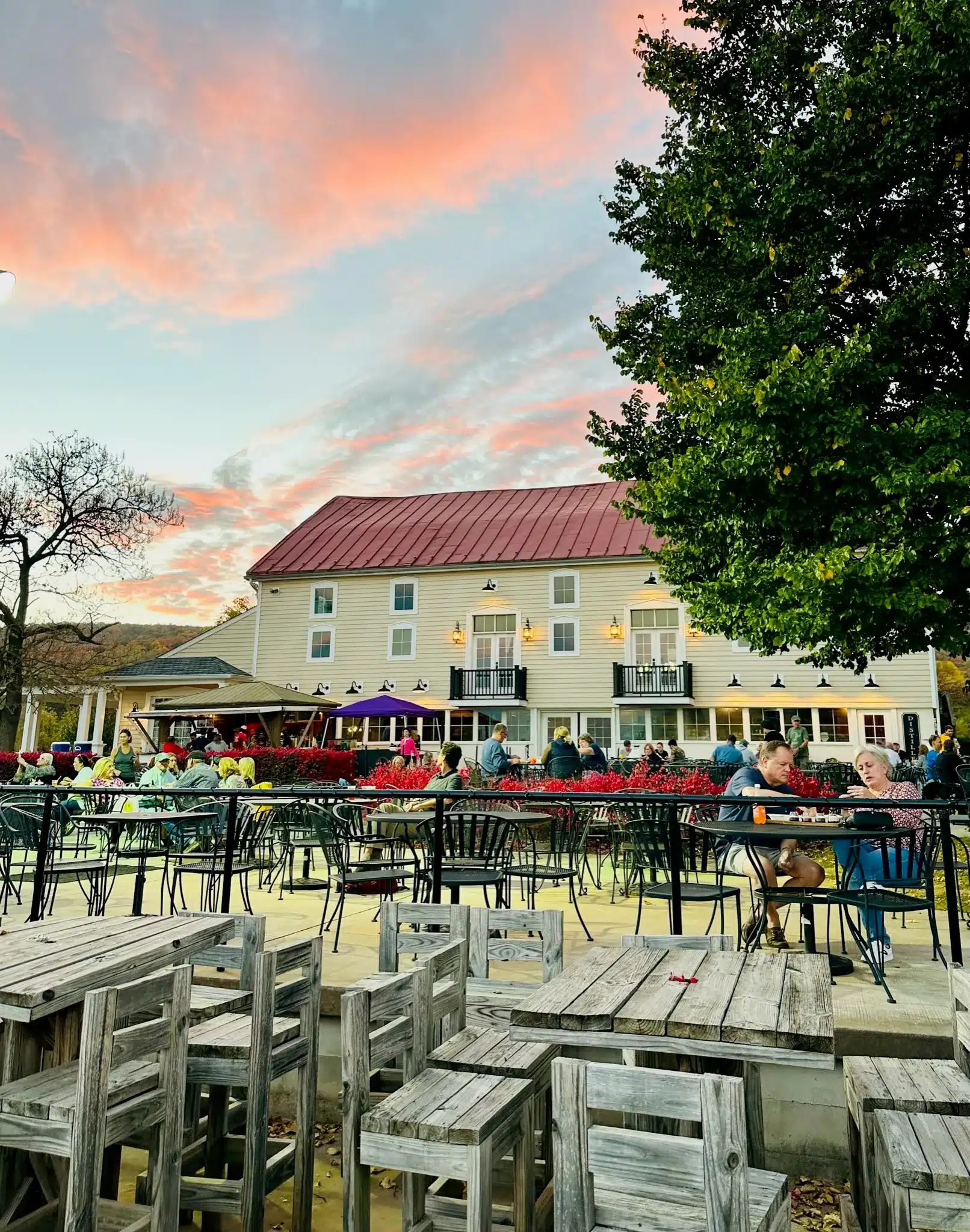 Outdoor Dining Area with Wooden Tables and Chairs Some Occupied by People Enjoying Meals and Conversation Nearby Greenery and a Large Beige Building Set the Scene Beneath a Colorful Sunsetperfect for Relaxing After Visiting Frederick Md Wineries