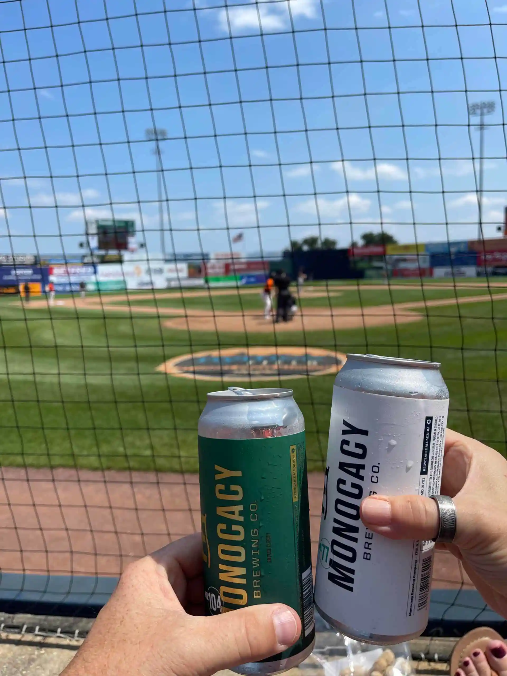 Two Hands Toast with Cans of Monocacy Brewing Co Beer at a Baseball Stadium Field and Players in View Under a Blue Skyperfect for Enjoying a Game Alongside the Frederick Keys Schedule