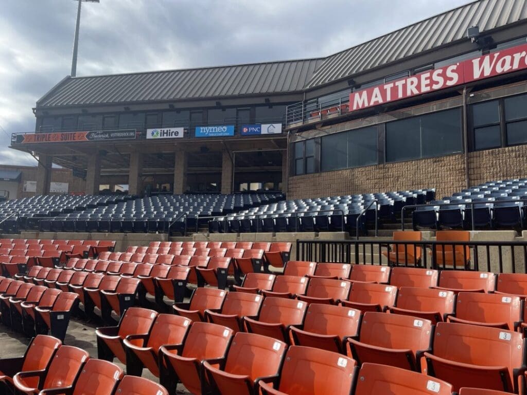 Rows of Empty Stadium Seats with Red Seats in the Foreground and Dark Blue Seats Behind a Covered Area with Sponsor Signs Including One Reading Mattress is Visible Above the Seatscatch It Live on the Frederick Keys Schedule the Sky is Cloudy