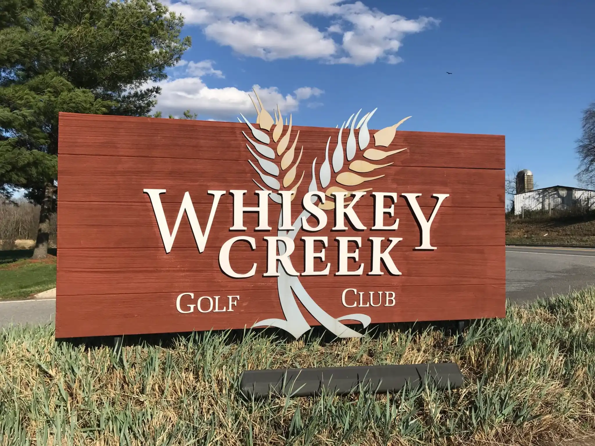 a Large Wooden Sign Reading Whiskey Creek Golf Club in White Letters with Two Stylized Wheat Stalks Crossing Behind the Text Stands on Grass Beside a Road in New Market Md Trees a Blue Sky with Clouds and Distant Farm Buildings Are in the Background