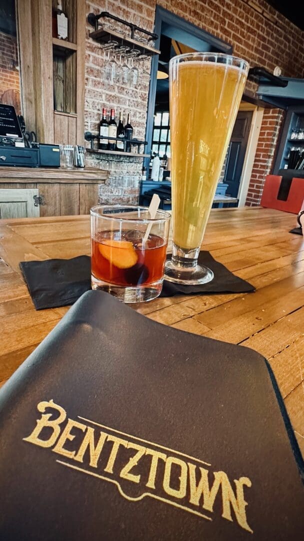 a Tall Light Beer and a Cocktail with an Orange Slice Sit on a Wooden Bar a Black Menu with Gold Bentztown Text is Visible Hinting at Why This Spot Ranks Among the Best Bars in Frederick Bottles and Exposed Brick Walls Create a Rustic Ambiance
