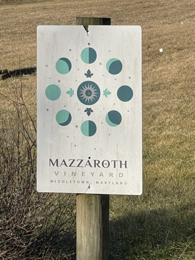 a Sign Attached to a Wooden Post Reads Mazzaroth Vineyard Middletown Marylanda Gem Among Frederick Md Wineries Above the Text Phases of the Moon Circle a Sun Like Symbol on a Weathered White Background Grass and Dried Bushes Frame the Scene