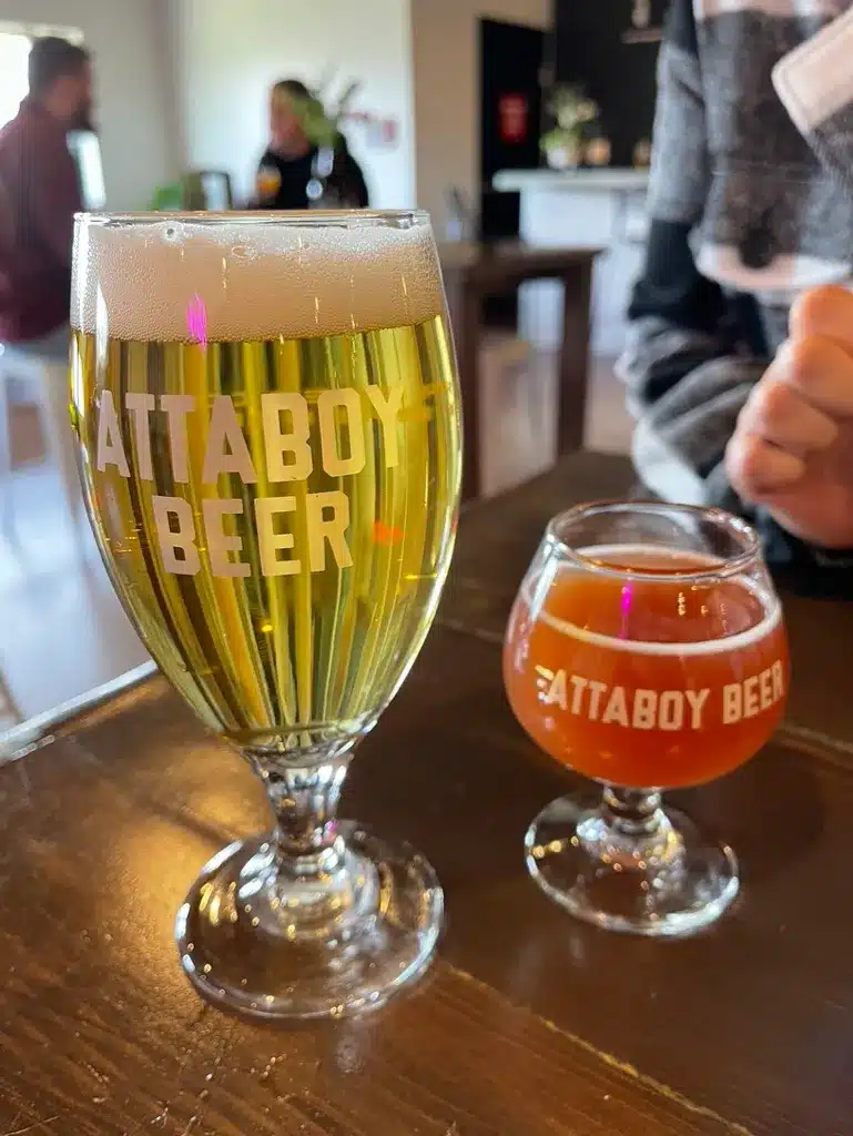 Two Glasses of Beer on a Wooden Table Feature Attaboy Beer Branding Showcasing the Vibrant Scene of Local Breweries Frederick Offers a Pale Golden Beer and an Amber Brew Highlight the Variety at One of the Top Breweries in Frederick Md
