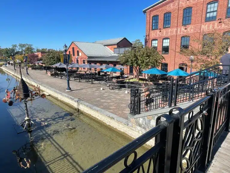 Outdoor Dining Area with Tables and Blue Umbrellas Lines a Brick Patio Next to a Calm Canal Steps Away Some of the Best Breweries in Frederick Md Offer Craft Brews Surrounded by Red Brick Buildings Green Trees and a Clear Blue Sky