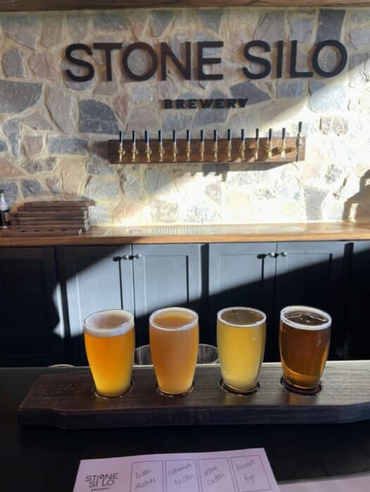 a Wooden Paddle Holds Four Small Glasses of Beer in Varying Shades on a Bar Counter Behind Them Stone Silo Brewery is Displayed Above Beer Taps Capturing the Inviting Atmosphere Found at the Best Breweries in Frederick Md Sunlight Casts Shadows Across the Scene