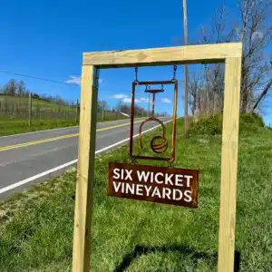 A wooden frame holds a metal sign for “Six Wicket Vineyards” by a rural roadside. Known among Frederick MD Wineries, the sign features a stylized emblem above the name, set on a grassy slope with fences, trees, and blue sky in the background.