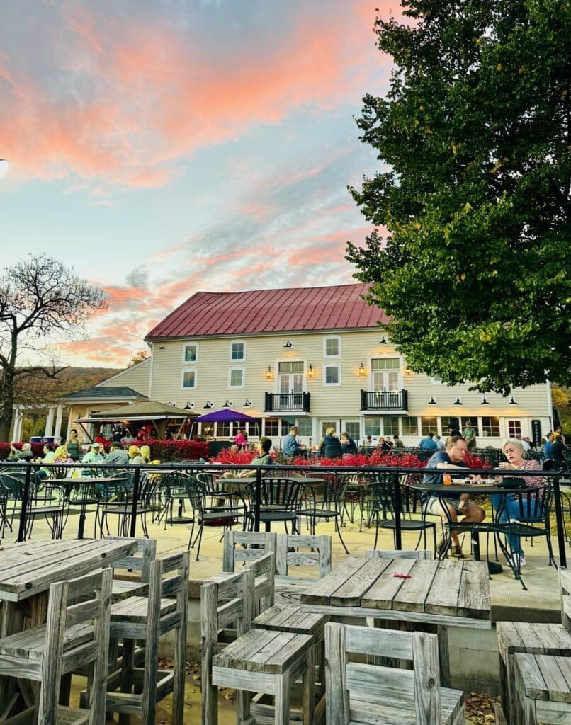 an Outdoor Dining Area with Wooden and Metal Tables and Chairs is Filled with People Socializing Behind Them Stands a Large Beige Building Similar to Some Hotels in Frederick Md Under a Colorful Sunset Sky Framed by Green Trees