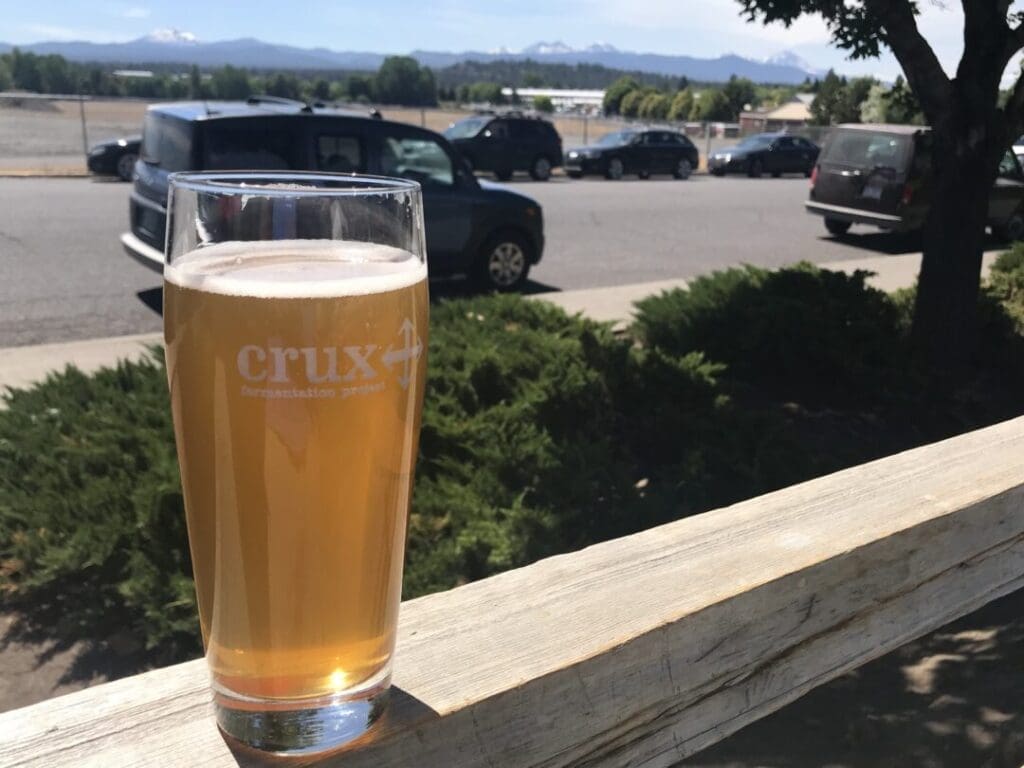 a Pint Glass of Hazy Golden Beer from One of the Best Breweries Bend Has to Offer Sits on a Wooden Railing Outdoors with Greenery Parked Cars and Snow Capped Mountains Beneath a Sunny Blue Sky in the Background