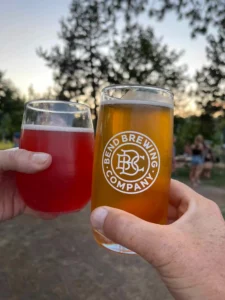Two hands clink glasses outdoors; one holds a red drink in a stemless wine glass, the other a pint of amber beer with the Bend Brewing Company logo. Trees and people blur in the background, evoking a casual dusk gathering in Bend, Oregon.